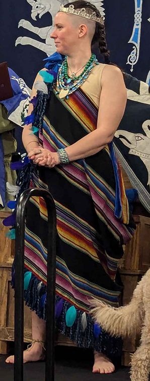 Tlatocapilli Z, one of the reigning Coronets of The Summits, wearing a black and rainbow tilmàtli decorated with blue and white feathers.