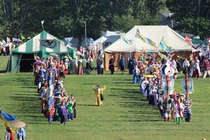 An overhead view of a field with tents on the far side. Between the viewer and the tents, two long processions approach the viewer, each with banners flying. In the large space between the processions, a single figure in a yellow dress stands with their back to the viewer.