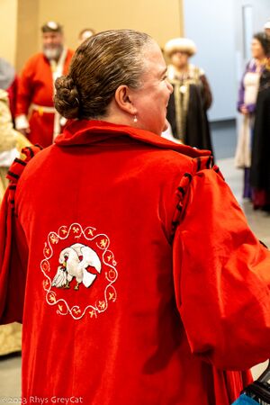 A white woman with auburn hair stands with her back to the viewer, her head turned so it can be seen in profile. Her hair is pulled back in a low bun, and she is smiling. She is wearing a high-collared red velveteen coat, the back of which is embroidered with a white pelican in its piety surrounded by a gold wreath.