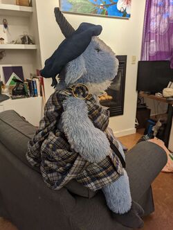 Bartholomew the Goat, a blue stuffed Easter goat, shown sitting on a grey armchair with a blue hat where his horns poke out, a blue plaid kilt with the ends wrapped over his shoulder and fastened with an annular cloak pin.