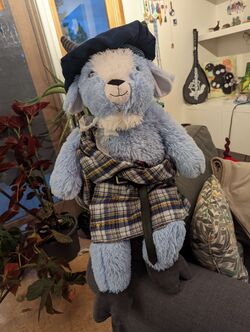 Bartholomew the Goat, a blue stuffed Easter goat, shown sitting on a grey armchair with a blue hat where his horns poke out, a blue plaid kilt with the ends wrapped over his shoulder and fastened with an annular cloak pin.